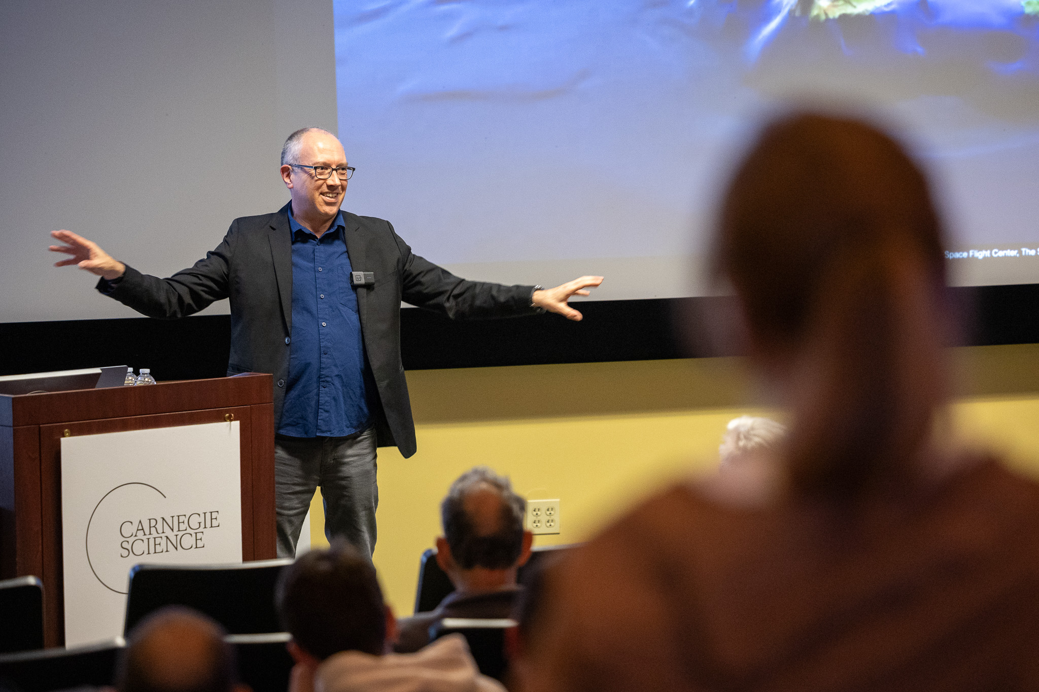 Caleb Scharf at NLS with Carnegie Science sign. Hands spread wide to describe the breadth of human possibility. 