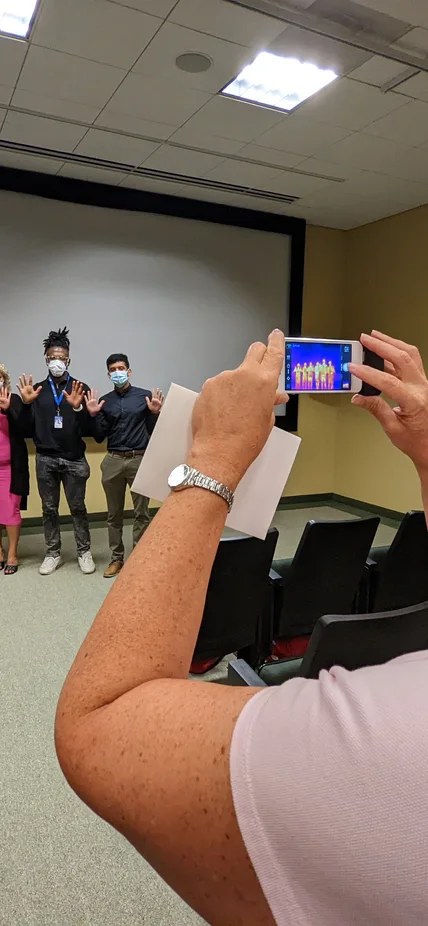 Alycia Weinberger holds up a phone to take a photo of a group of six interns. The phone screen is visible and the image is showing the infrared heat map of the interns.