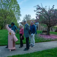 Scientists and members of the public peer through a pastel pink telescope. 