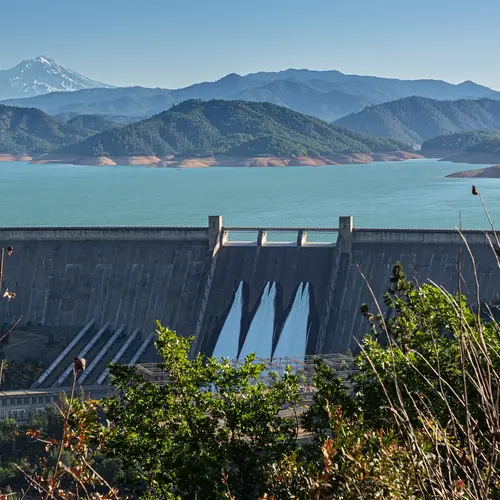 Shasta Dam in Northern California as seen from a distance. Mountains are green in the background. 